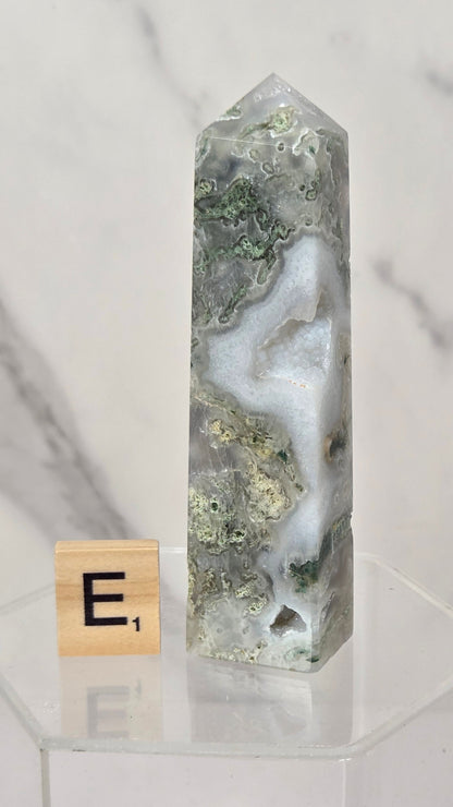 A polished agate crystal tower with natural green and white marbling, displayed on a clear acrylic stand next to a wooden Scrabble tile showing the letter E.
