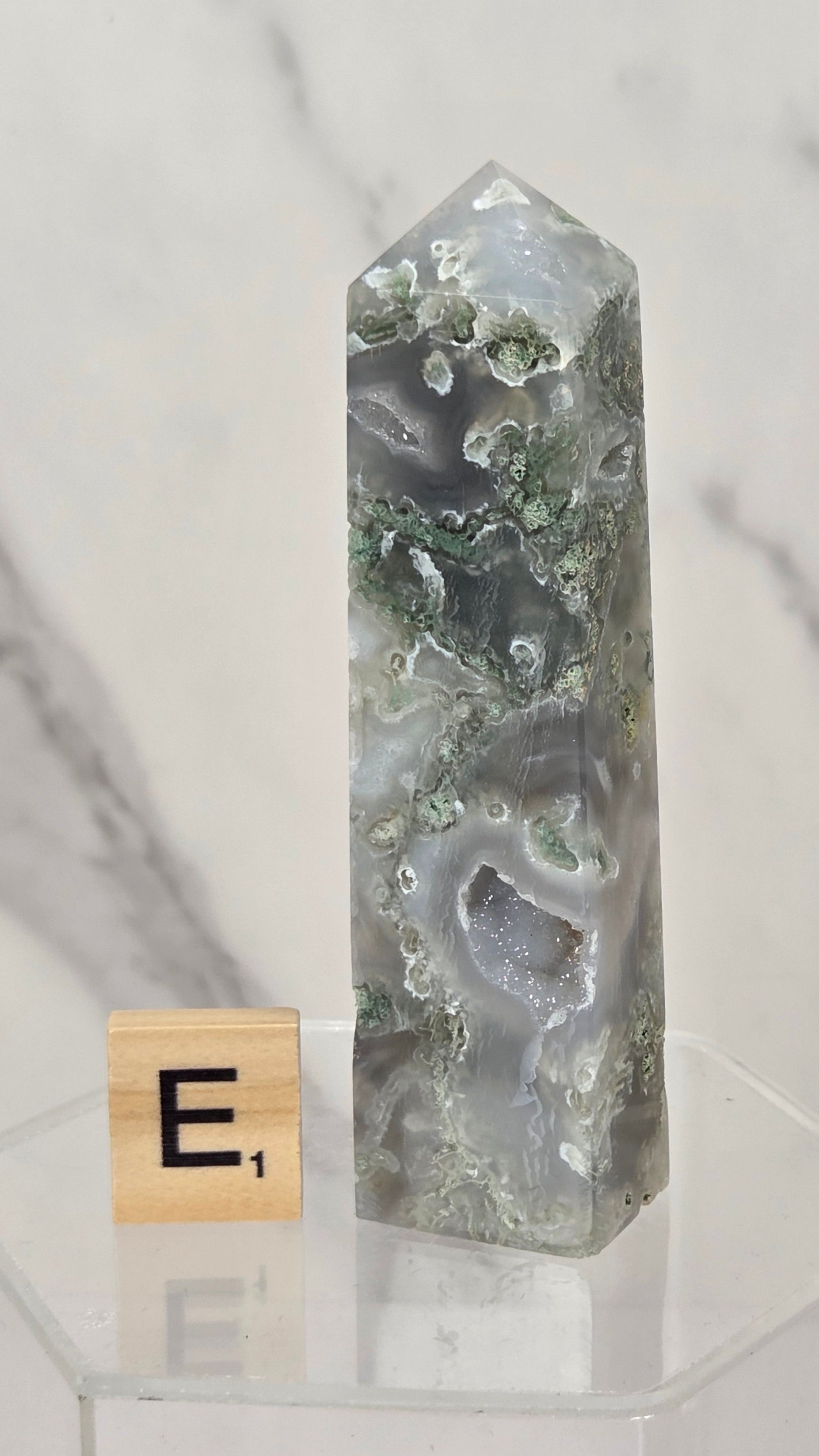 A polished agate crystal tower with natural green and gray banded patterns, displayed on a clear acrylic stand next to a small Scrabble tile showing the letter 'E'.