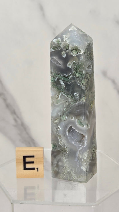 A polished agate crystal tower with natural green and gray banded patterns, displayed on a clear acrylic stand next to a small Scrabble tile showing the letter 'E'.