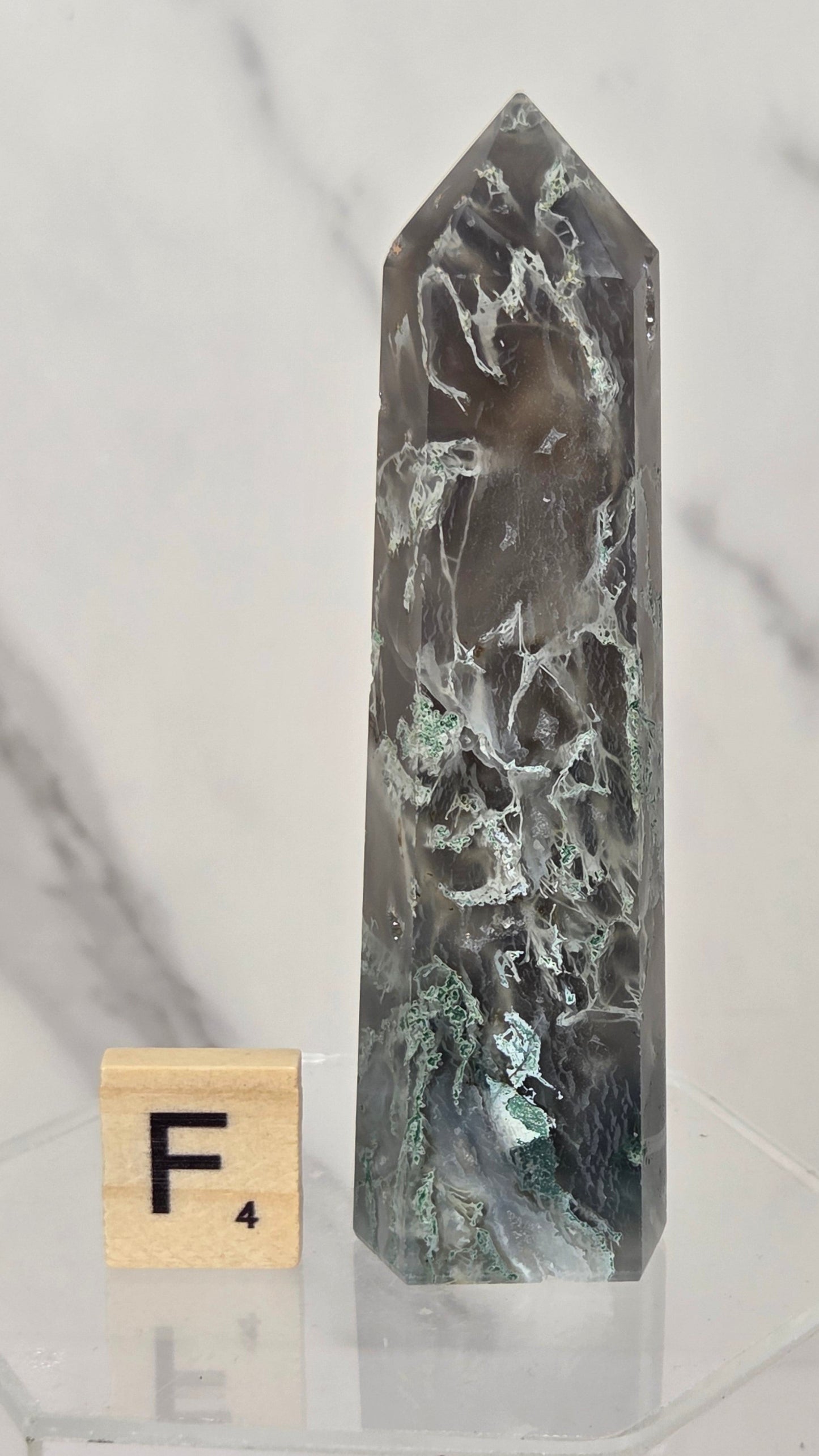 A tall, pointed crystal tower with a mottled gray, black, and green marbled pattern, displayed on a clear acrylic hexagonal base next to a wooden Scrabble tile showing the letter F.