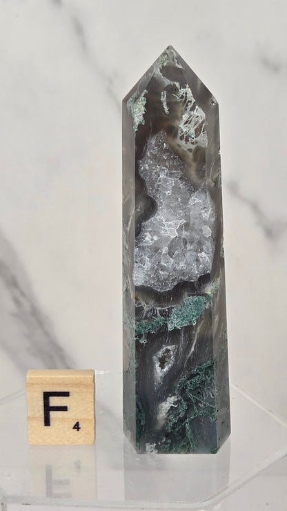 A polished crystal point specimen with natural marbled patterns in gray, black, and green, displayed on a clear acrylic stand next to a wooden Scrabble tile showing the letter F.