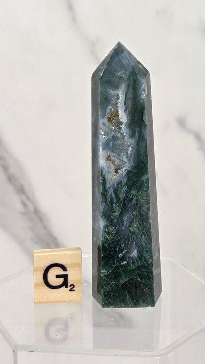 A polished dark green and blue geode point crystal tower with natural inclusions, displayed on a clear acrylic stand next to a wooden Scrabble tile showing the letter 'G' on a white marble background.