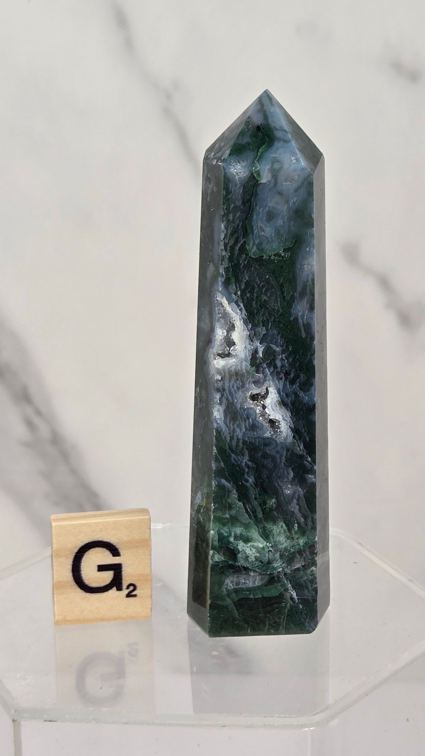 A polished dark green and black crystal point tower with natural marbling, displayed on a clear acrylic stand next to a wooden Scrabble tile showing the letter G.