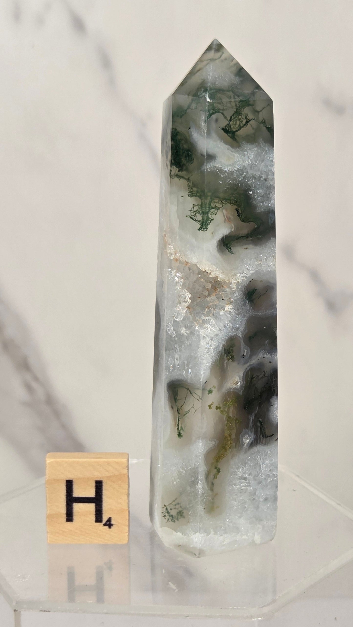 A tall, pointed clear quartz crystal point with natural white, gray, and dark green agate inclusions, displayed on a clear acrylic stand next to a small Scrabble tile showing the letter 'H'.