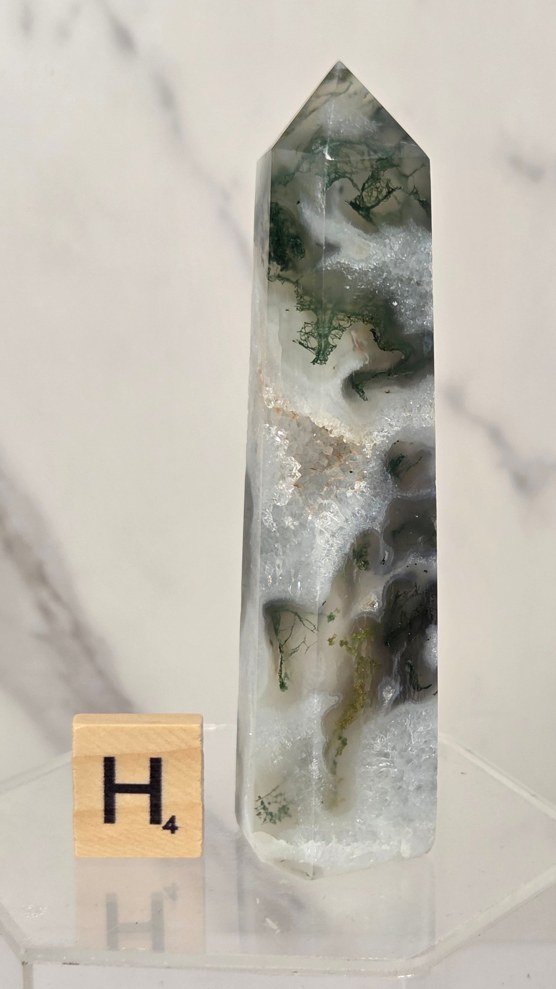 A tall, pointed clear quartz crystal point with natural white, gray, and dark green agate inclusions, displayed on a clear acrylic stand next to a small Scrabble tile showing the letter 'H'.