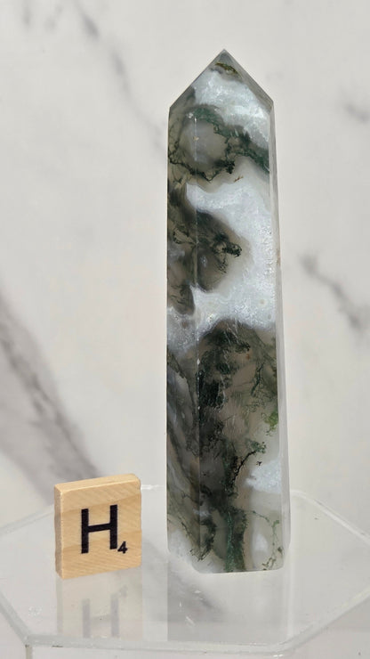 A tall clear crystal point with dark green and cloudy white inclusions, displayed on a clear acrylic stand next to a wooden Scrabble tile showing the letter H.