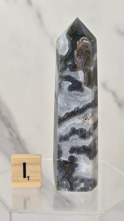 A tall, polished agate crystal point with natural wavy bands of gray, white, and dark charcoal, displayed on a clear acrylic stand next to a small wooden block with the number 1.