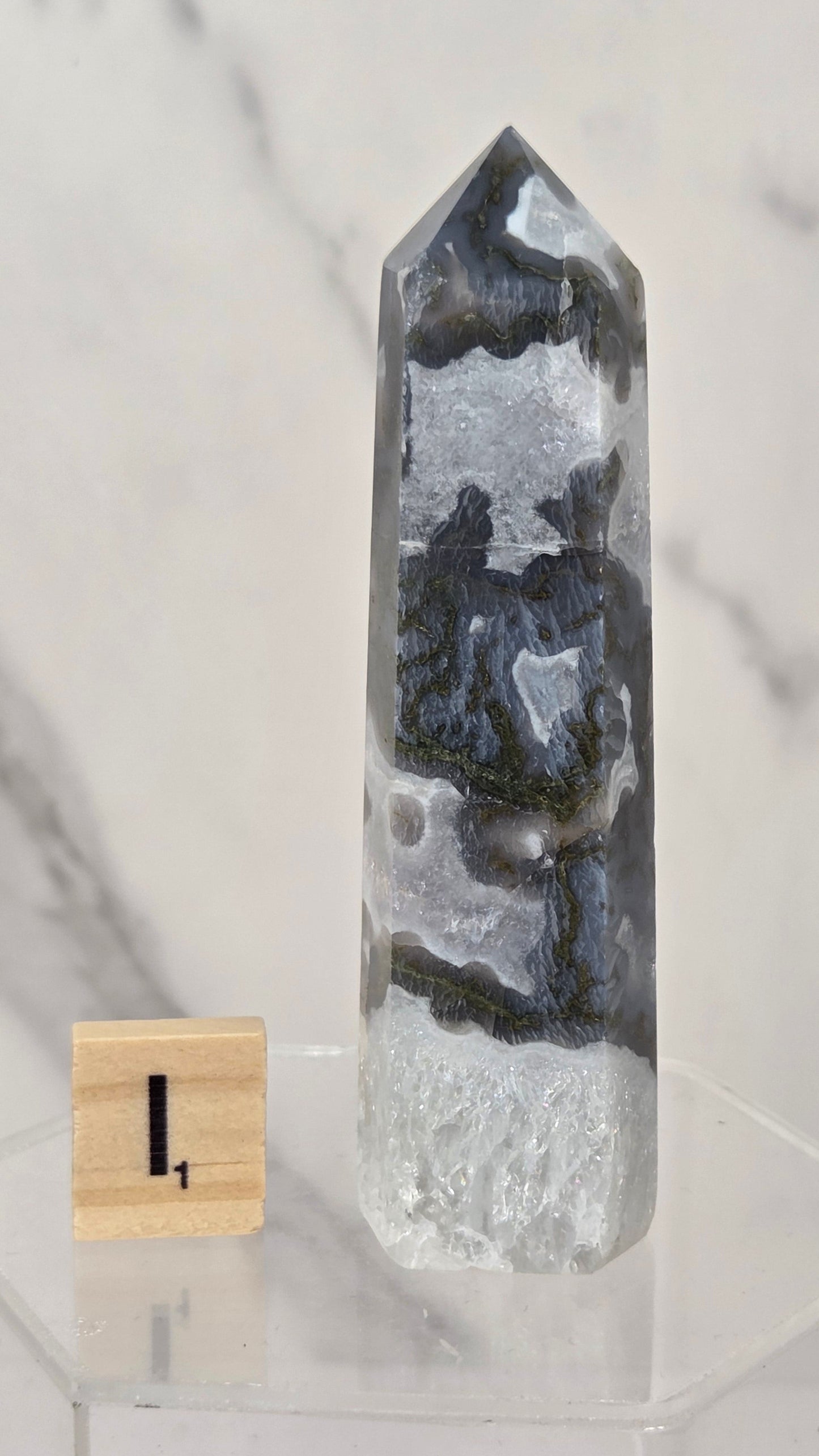 A tall pointed crystal tower with layered black, gray, and white agate-like patterns, displayed on a clear acrylic stand next to a small wooden block marked 1, against a light marble-patterned background.