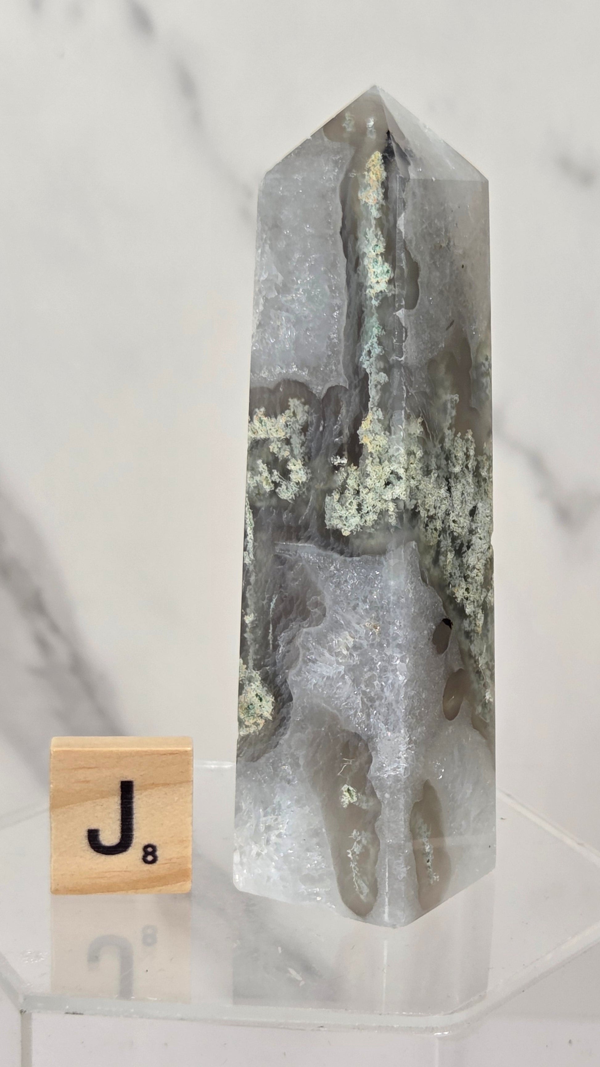 A tall, polished gray agate crystal point with natural veining and pale greenish inclusions, displayed on a clear acrylic stand next to a small wooden Scrabble tile with the letter J.