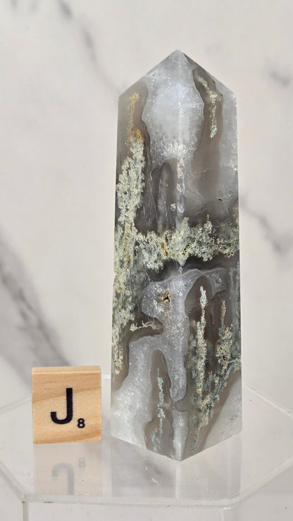 A tall, pointed agate crystal tower with natural banding in gray, white, and brown, displayed on a clear acrylic stand next to a wooden Jenga block on a white marble surface.