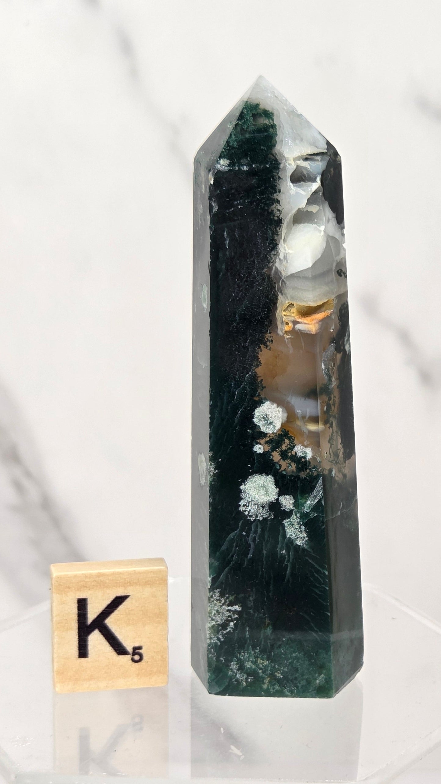 A tall, pointed clear crystal tower with dark green and black internal inclusions and small white floral details, displayed on a clear acrylic base next to a wooden Scrabble tile showing the letter K.