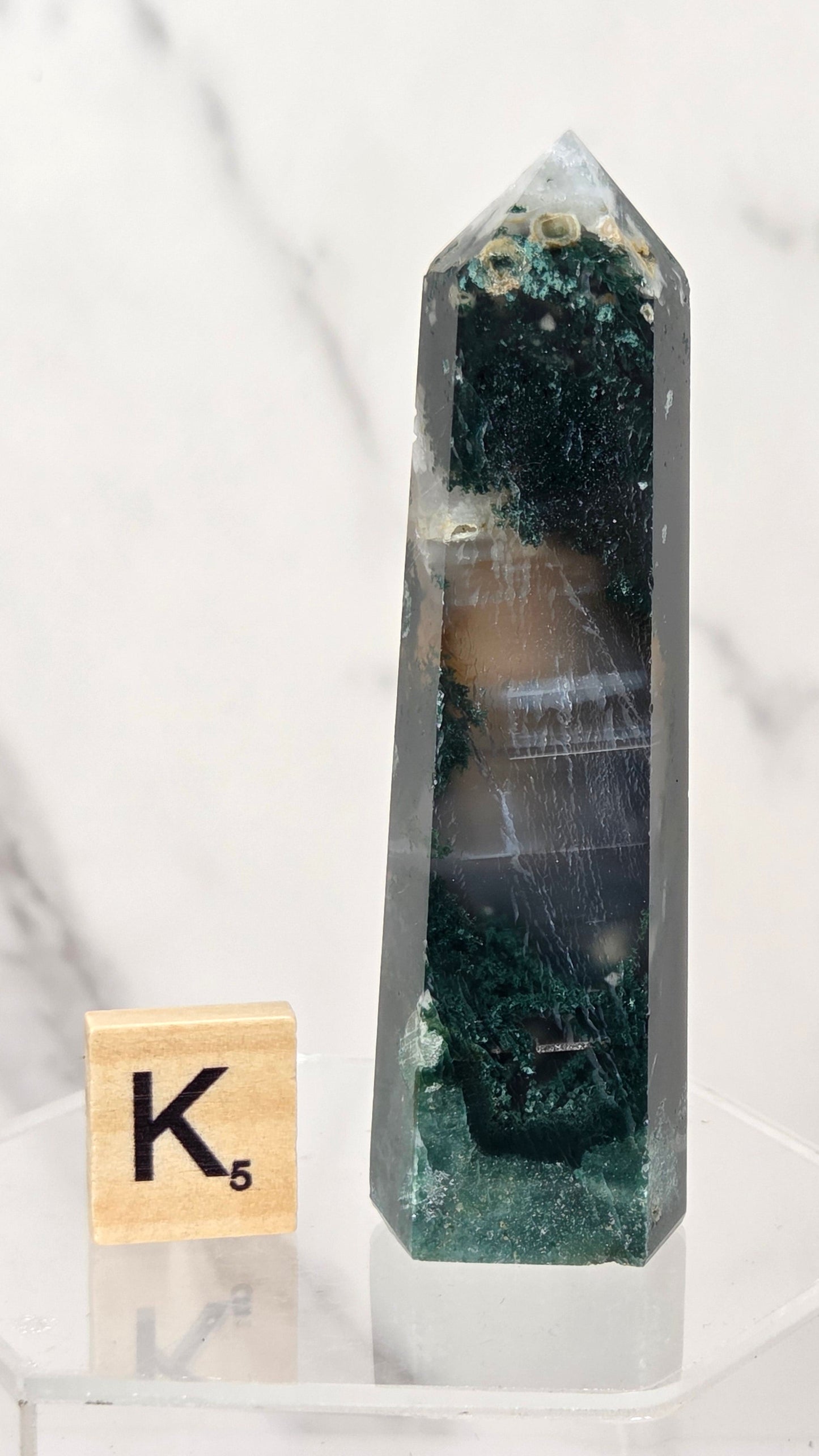 A tall, pointed clear quartz crystal tower with layered bands of dark green, black, and beige, displayed on a clear acrylic stand next to a wooden Scrabble tile showing the letter K.