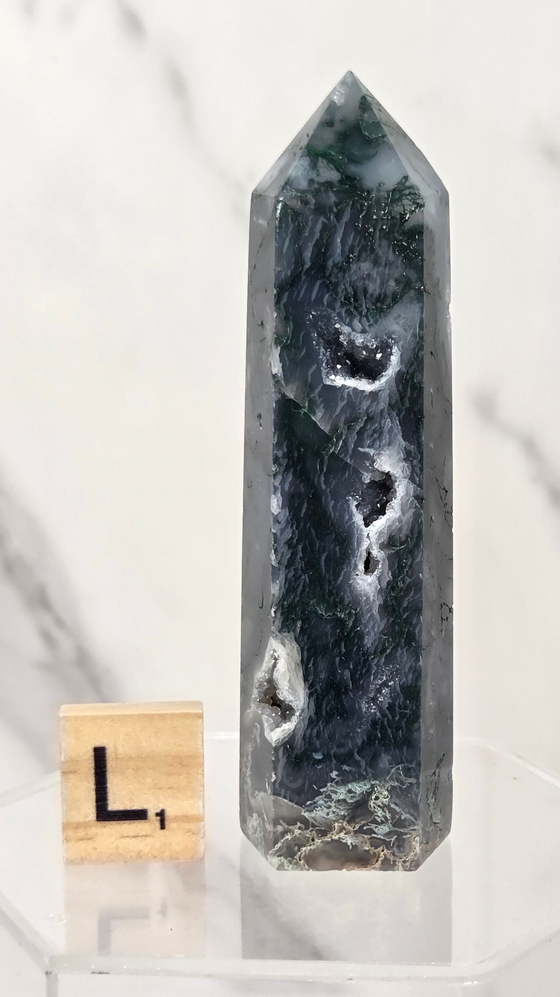 A tall polished natural crystal point with dark green, black, and white agate-like patterns, displayed on a clear acrylic stand next to a small wooden letter block.