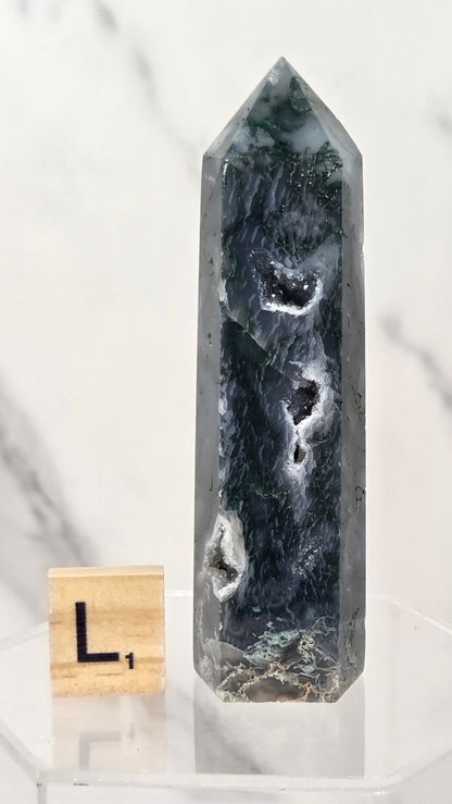 A tall polished natural crystal point with dark green, black, and white agate-like patterns, displayed on a clear acrylic stand next to a small wooden letter block.
