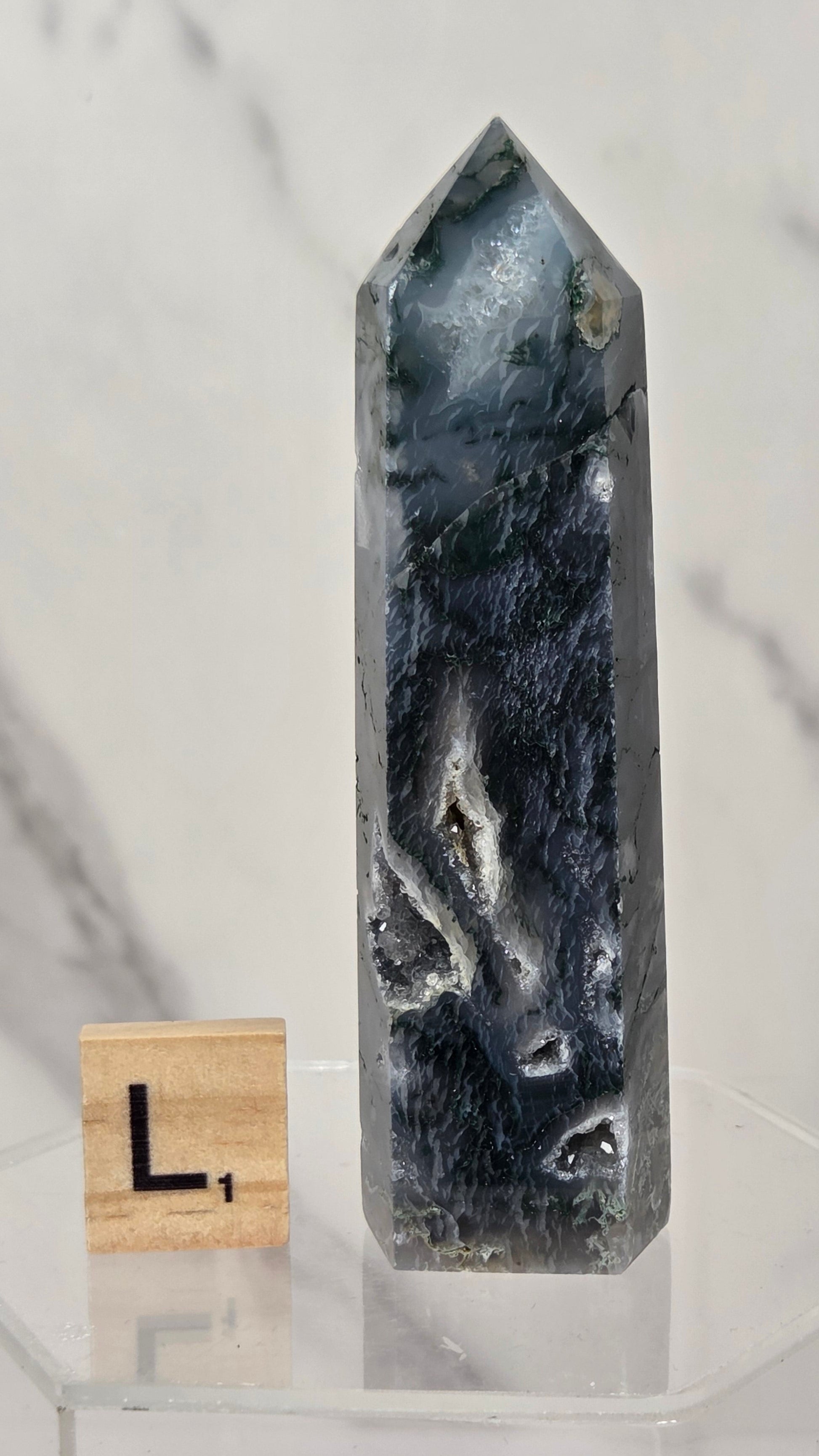 A tall, polished dark blue-gray crystal point displayed on a clear acrylic stand, with a small wooden letter block showing the letter L beside it, against a light marble-patterned background.
