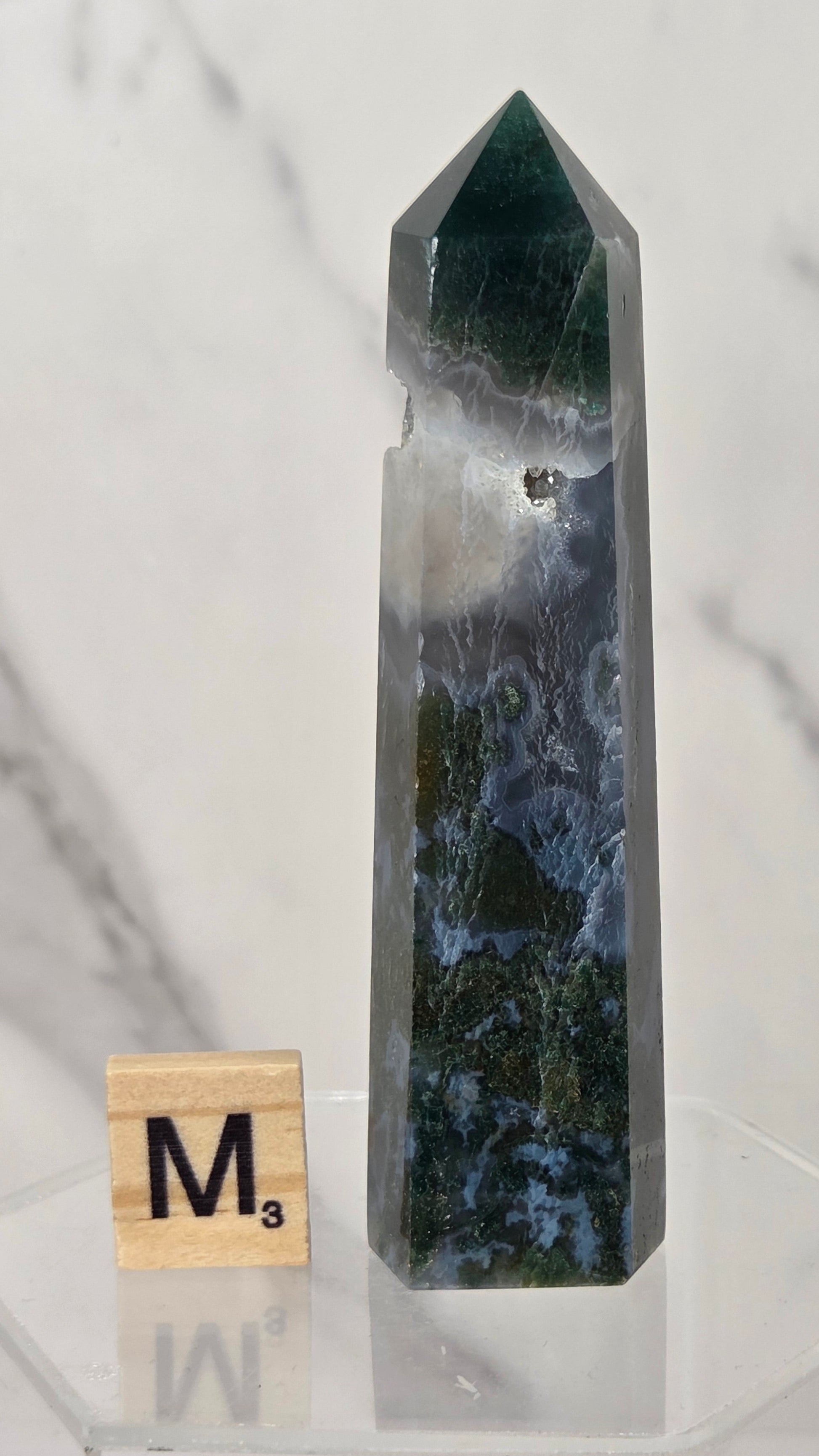 A polished natural crystal point with a marbled pattern of dark green, white, and blue, displayed on a clear acrylic stand next to a wooden Scrabble tile.