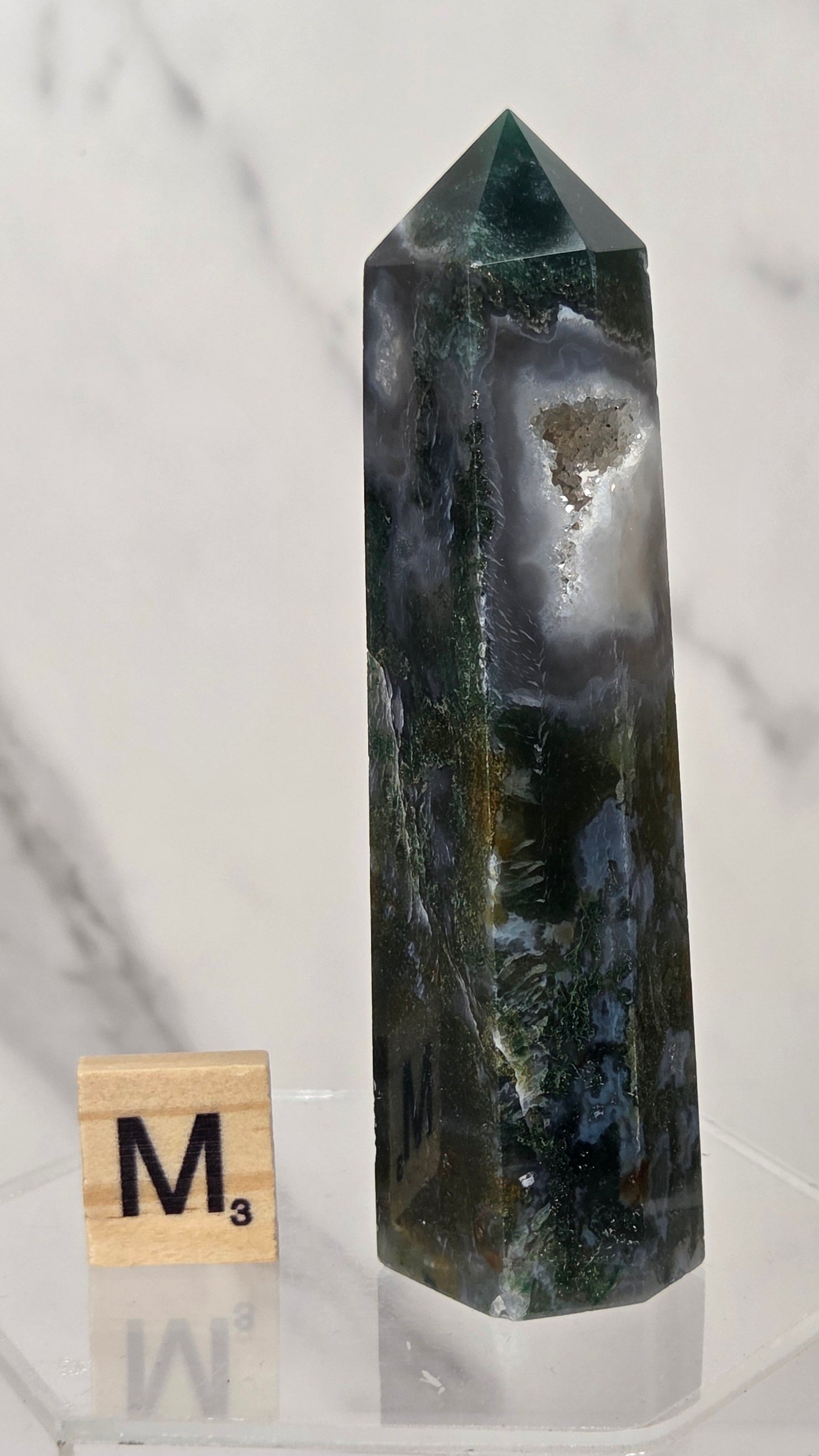 A dark green and black natural crystal tower with white and gray agate bands, displayed on a clear acrylic stand next to a wooden Scrabble tile marked M3.