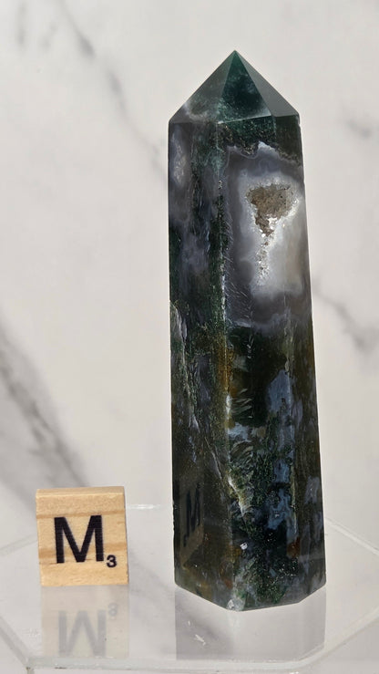 A dark green and black natural crystal tower with white and gray agate bands, displayed on a clear acrylic stand next to a wooden Scrabble tile marked M3.