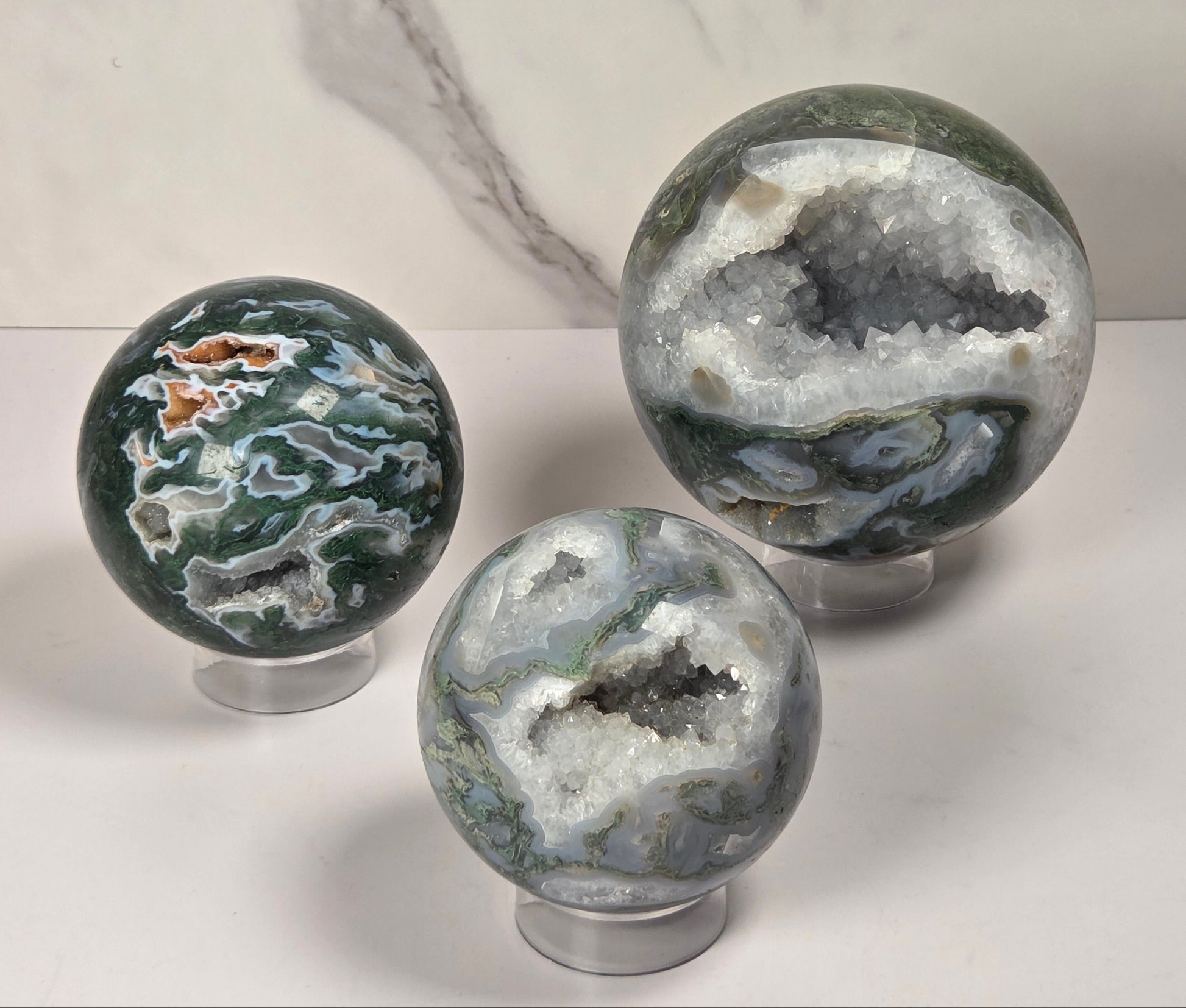 A set of polished agate crystal sphere with swirling green, gray, and black bands, displayed on a clear acrylic stand next to a wooden letter tile spelling 'A1' on a light marble surface.