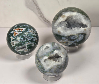 A set of polished agate crystal sphere with swirling green, gray, and black bands, displayed on a clear acrylic stand next to a wooden letter tile spelling 'A1' on a light marble surface.