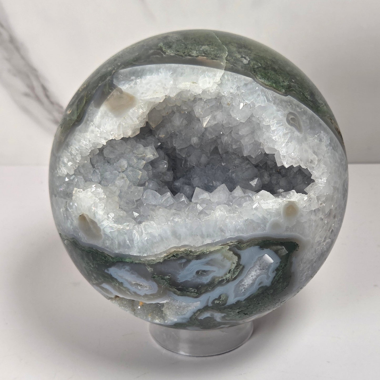 A polished agate crystal sphere with swirling green, gray, and black bands, displayed on a clear acrylic stand next to a wooden letter tile spelling 'A1' on a light marble surface.