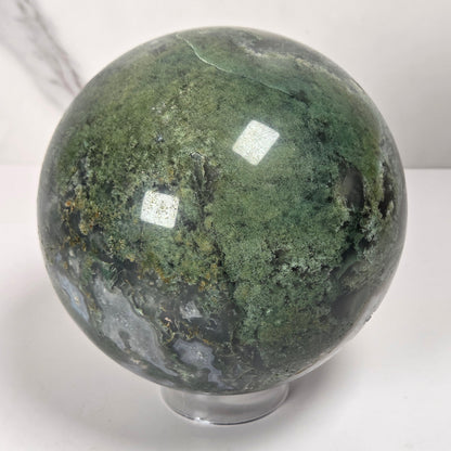 A polished agate crystal sphere with swirling green, gray, and black bands, displayed on a clear acrylic stand next to a wooden letter tile spelling 'A1' on a light marble surface.