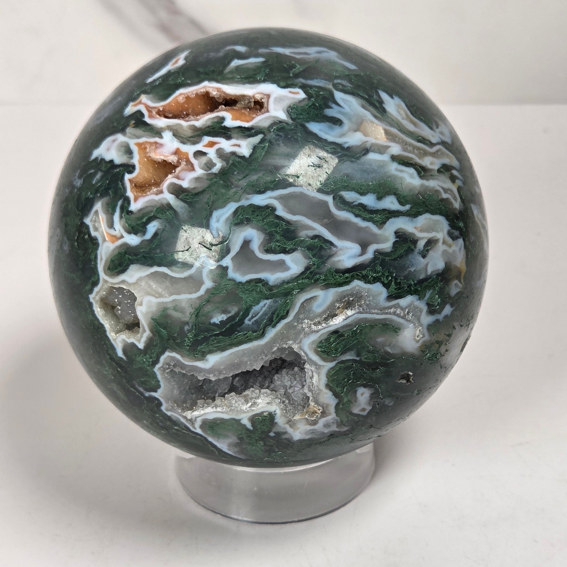 A polished agate crystal sphere with swirling green, gray, and black bands, displayed on a clear acrylic stand next to a wooden letter tile spelling 'A1' on a light marble surface.