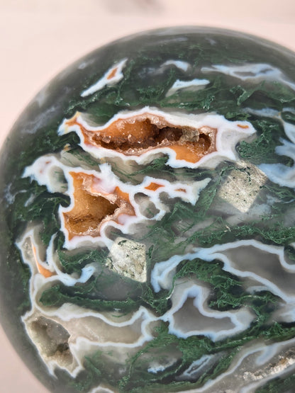 A polished agate crystal sphere with swirling green, gray, and black bands, displayed on a clear acrylic stand next to a wooden letter tile spelling 'A1' on a light marble surface.