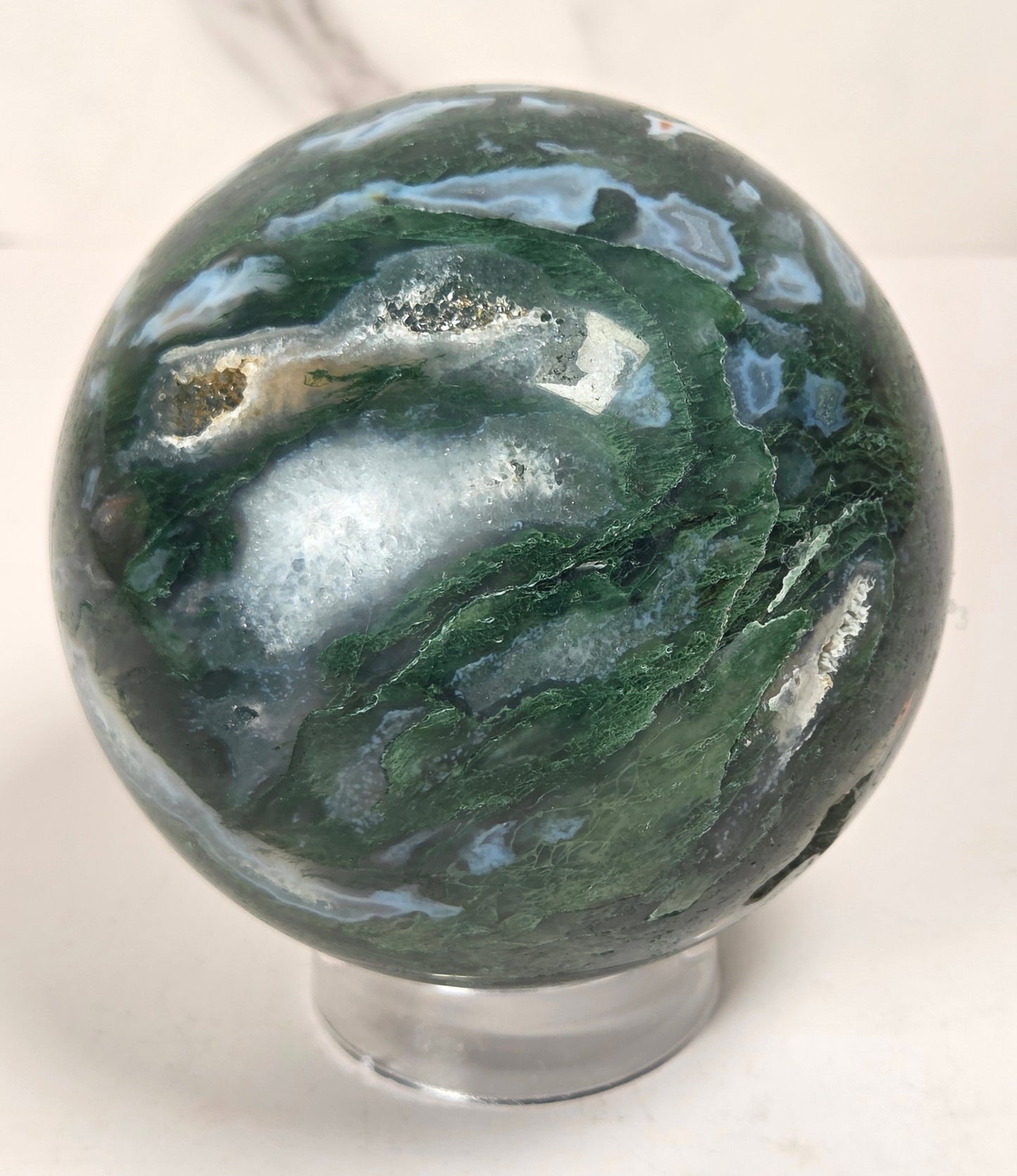 A polished agate crystal sphere with swirling green, gray, and black bands, displayed on a clear acrylic stand next to a wooden letter tile spelling 'A1' on a light marble surface.