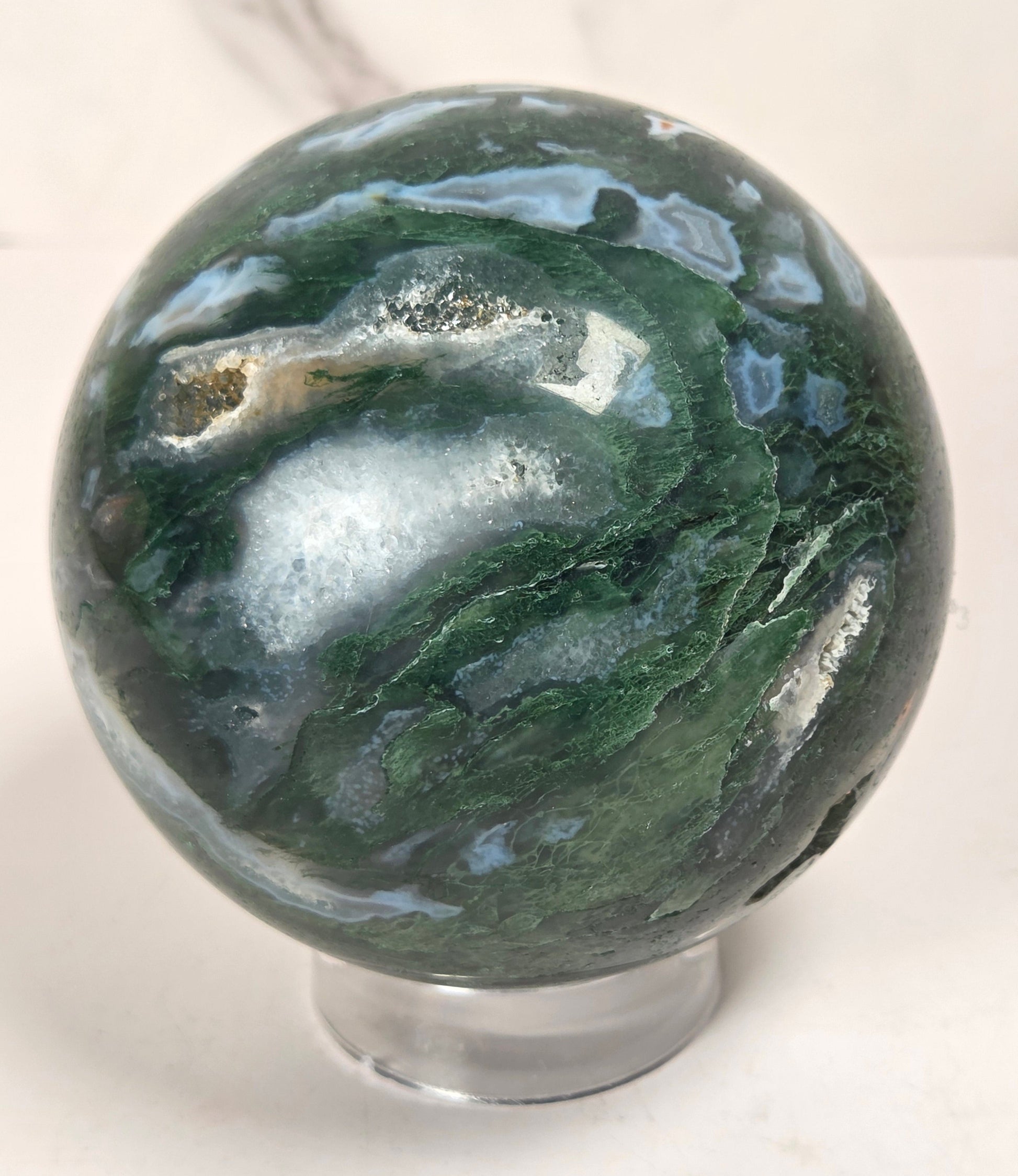 A polished agate crystal sphere with swirling green, gray, and black bands, displayed on a clear acrylic stand next to a wooden letter tile spelling 'A1' on a light marble surface.
