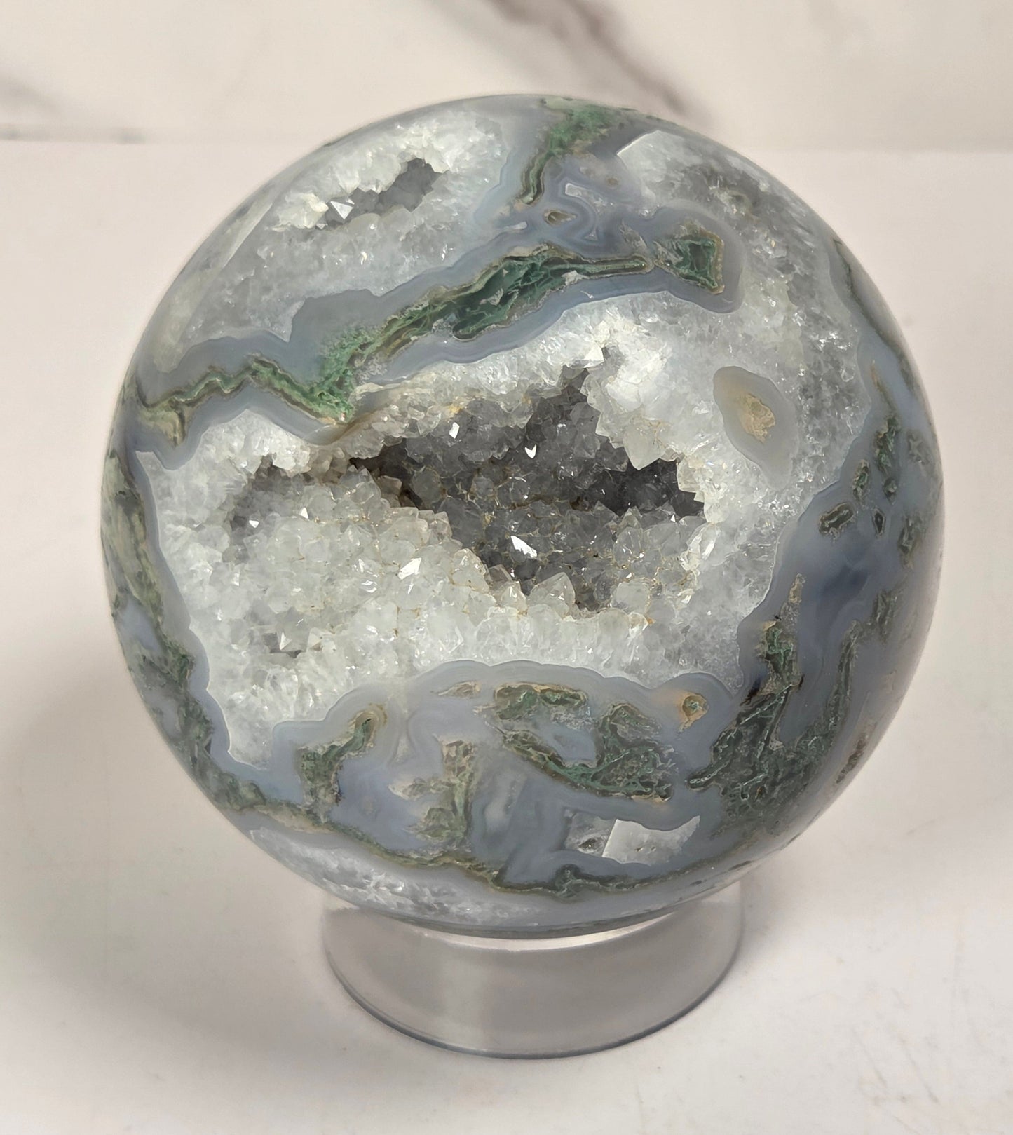 A polished agate crystal sphere with swirling green, gray, and black bands, displayed on a clear acrylic stand next to a wooden letter tile spelling 'A1' on a light marble surface.