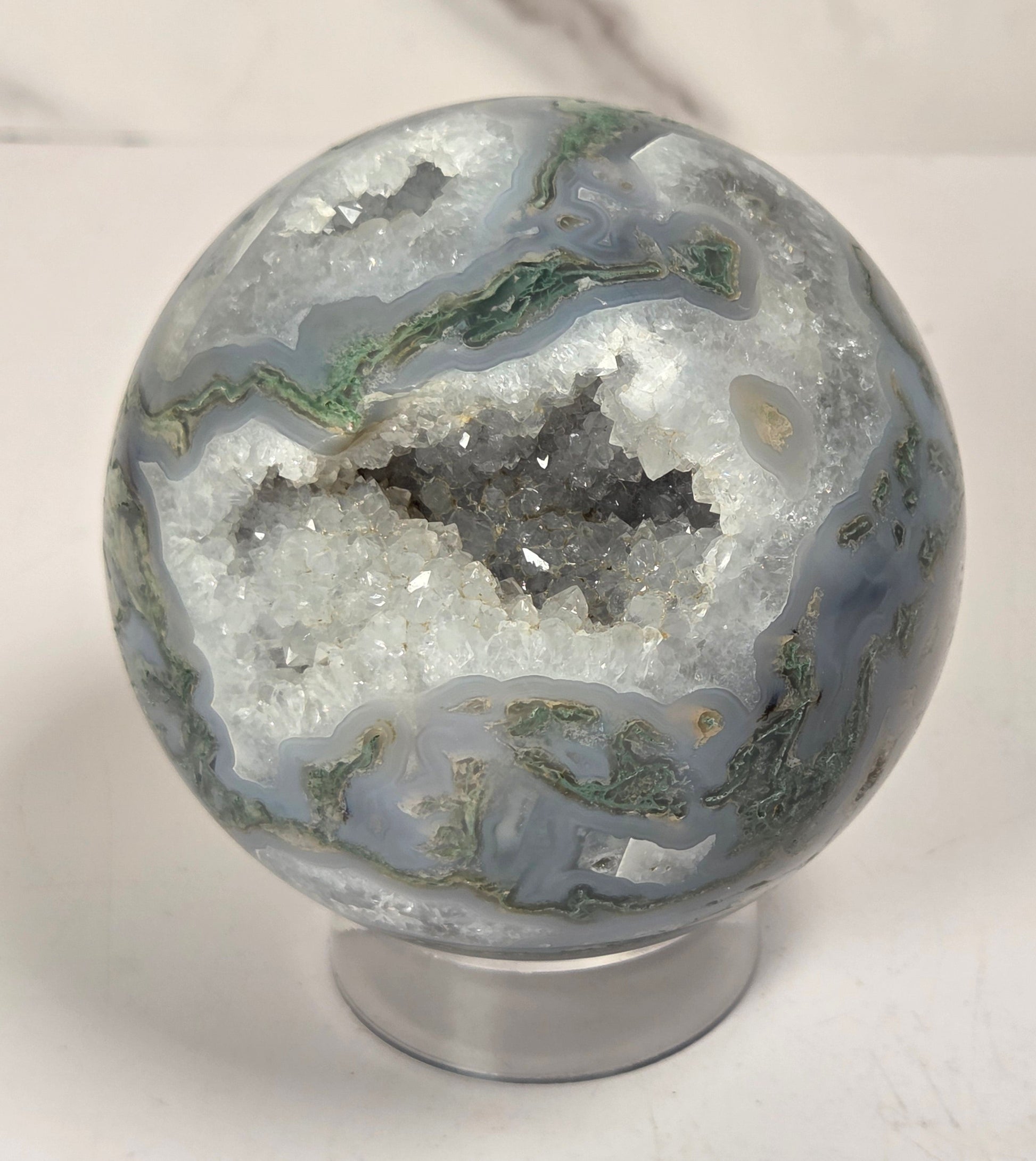 A polished agate crystal sphere with swirling green, gray, and black bands, displayed on a clear acrylic stand next to a wooden letter tile spelling 'A1' on a light marble surface.