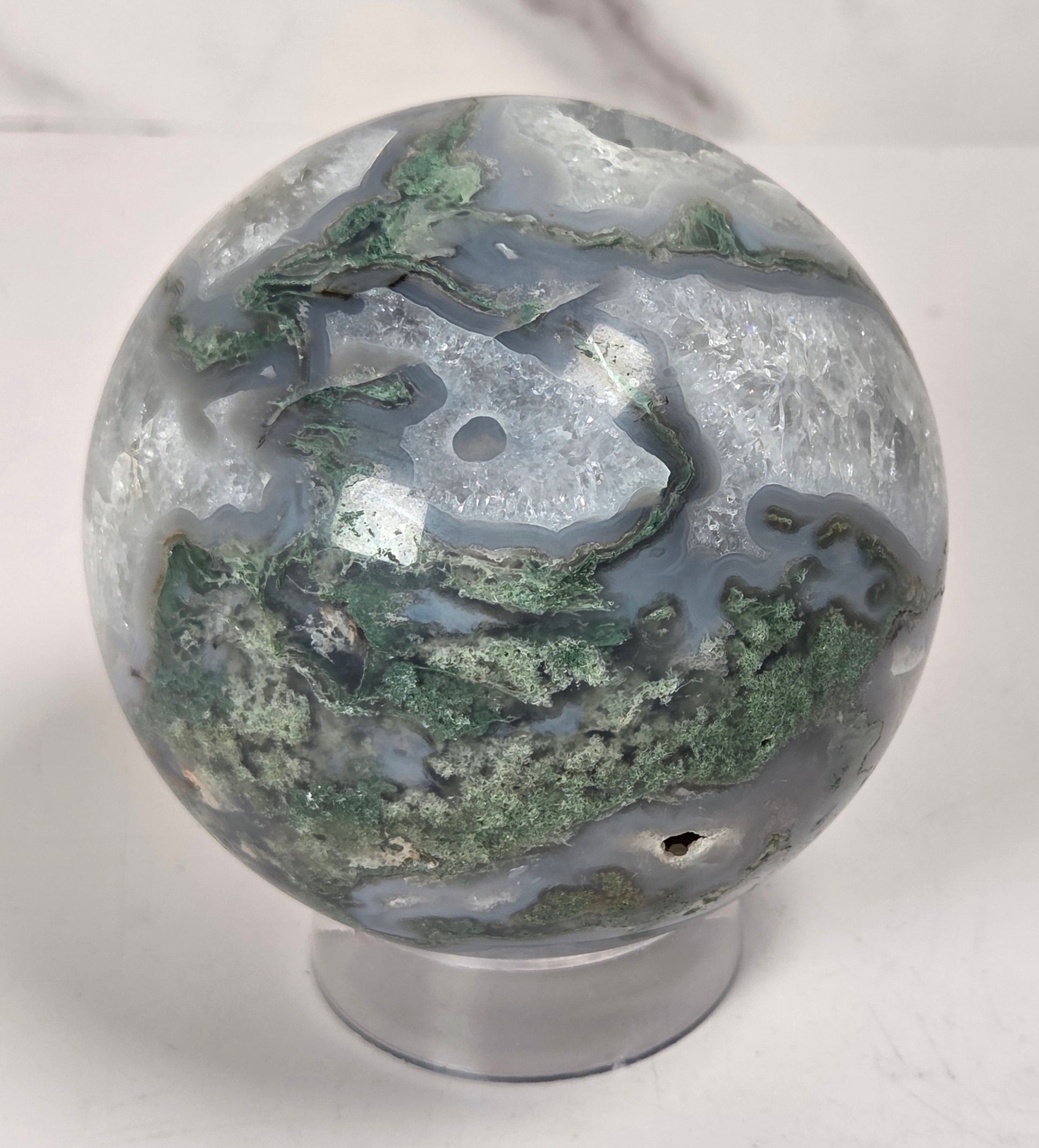 A polished agate crystal sphere with swirling green, gray, and black bands, displayed on a clear acrylic stand next to a wooden letter tile spelling 'A1' on a light marble surface.