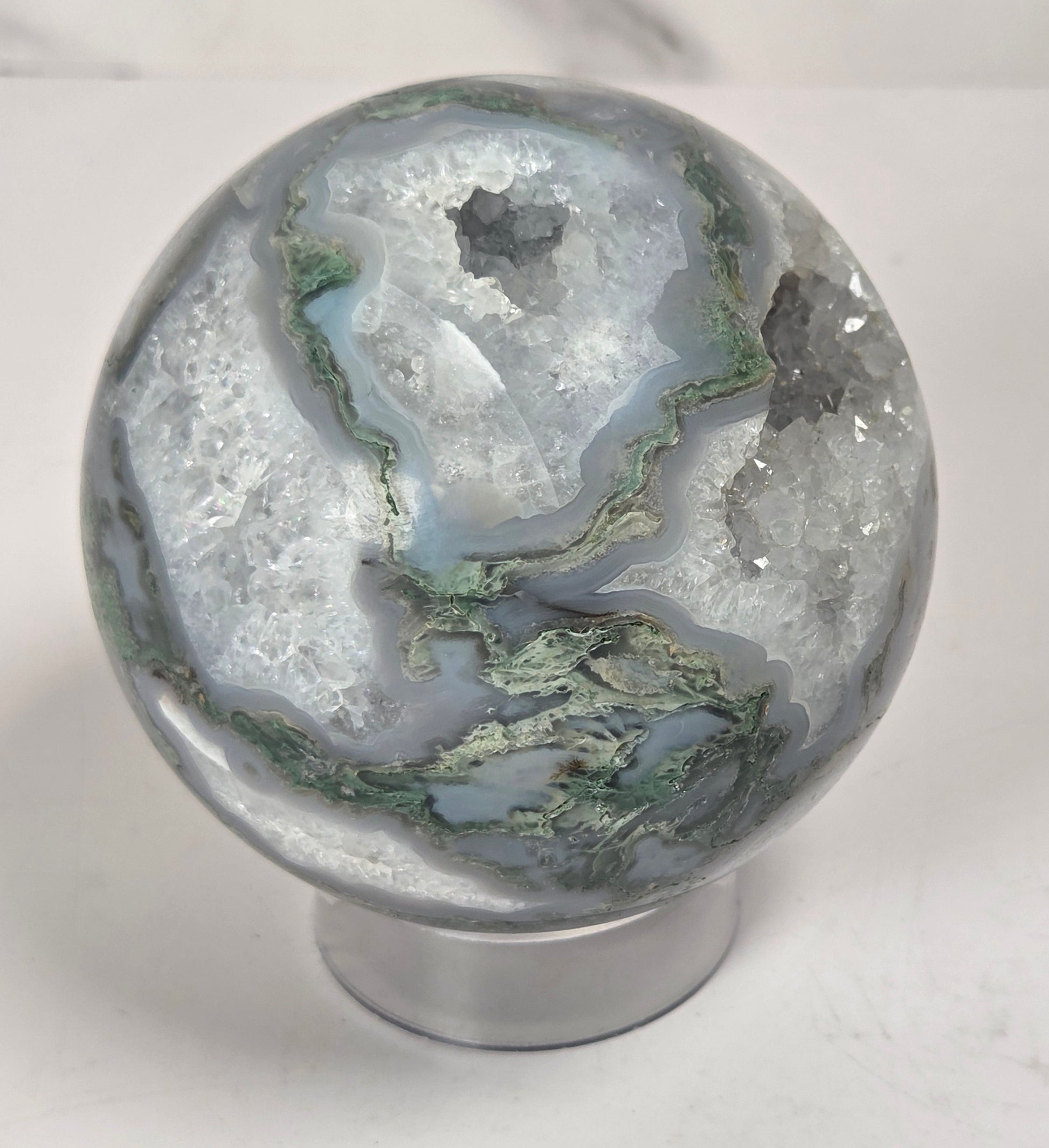 A polished agate crystal sphere with swirling green, gray, and black bands, displayed on a clear acrylic stand next to a wooden letter tile spelling 'A1' on a light marble surface.