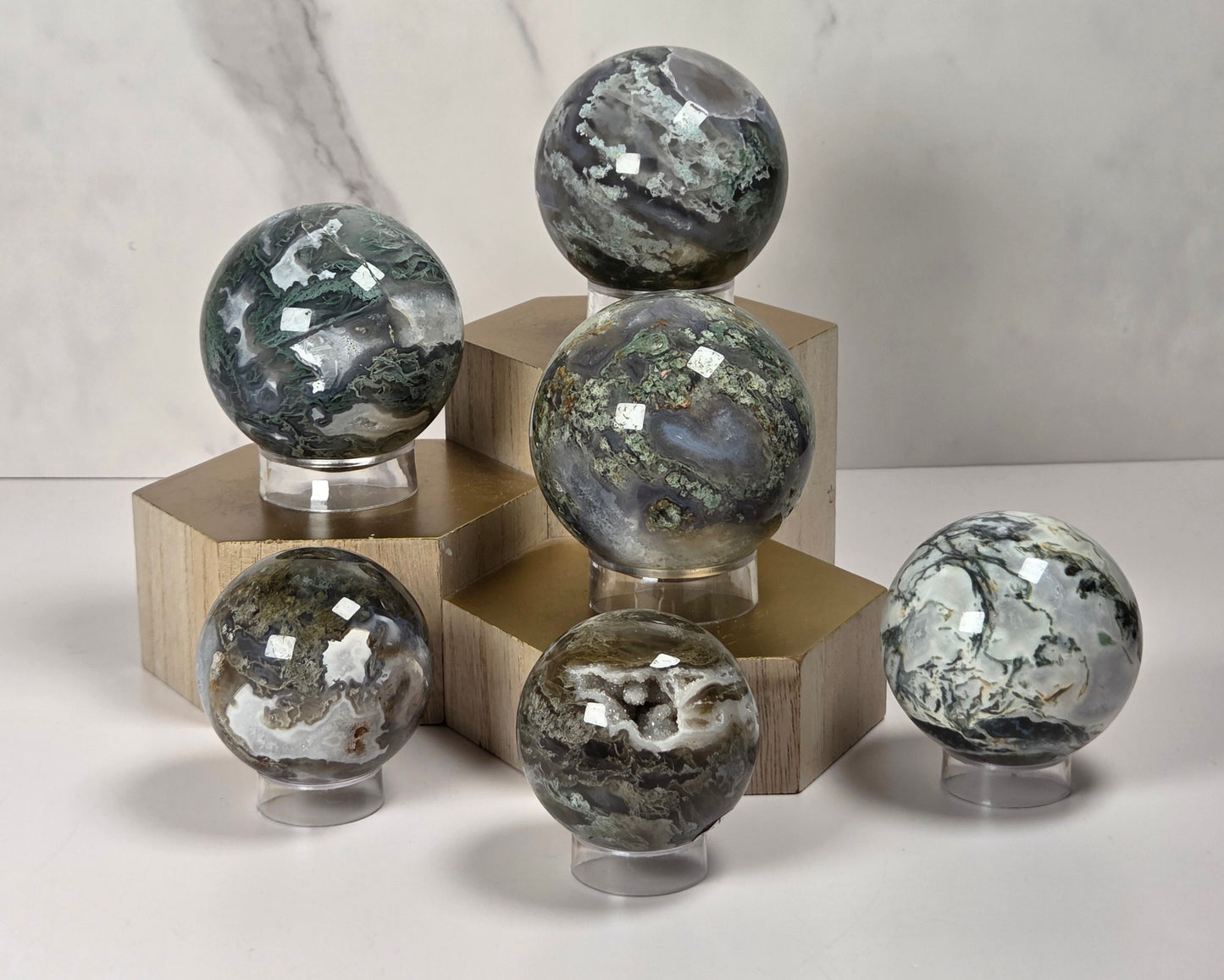 Six polished crystal spheres with natural stone patterns in shades of gray, green, and white, displayed on clear stands and wooden blocks against a light marble background.