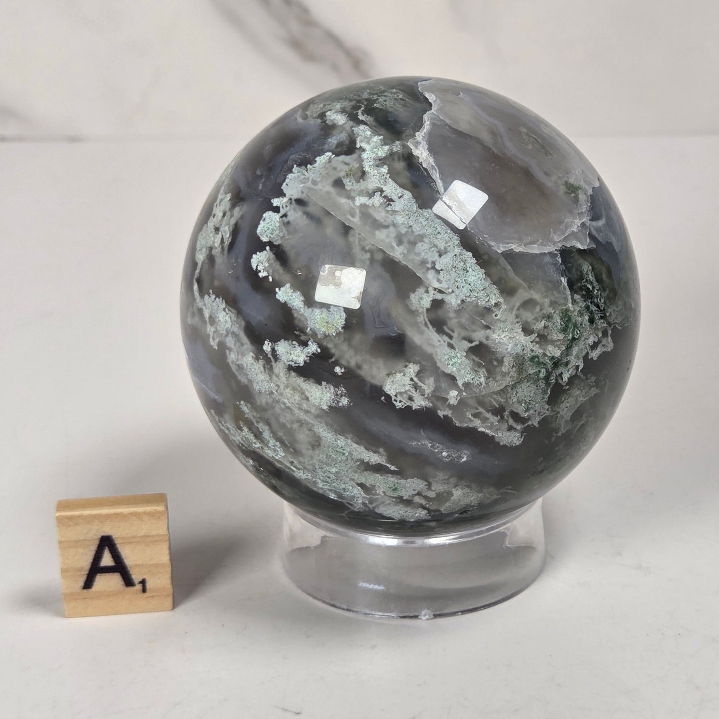 A polished gray agate crystal sphere with white and greenish veining, displayed on a clear acrylic stand next to a wooden letter tile showing the letter 'A', set against a light gray marble-patterned background.