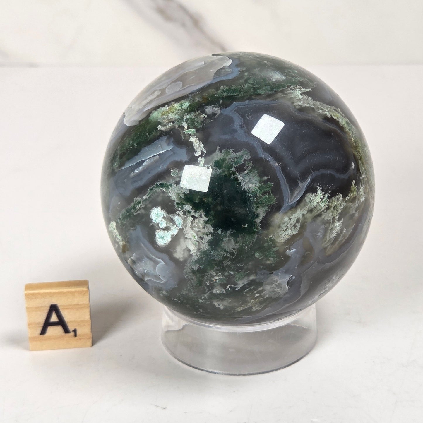 A polished agate crystal sphere with swirling green, gray, and black bands, displayed on a clear acrylic stand next to a wooden letter tile spelling 'A1' on a light marble surface.