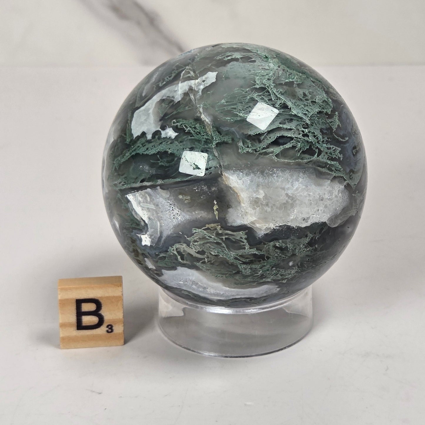 A polished round stone sphere with green, white, and gray marbled patterns, displayed on a clear acrylic stand next to a small wooden Scrabble tile showing the letter 'B'.