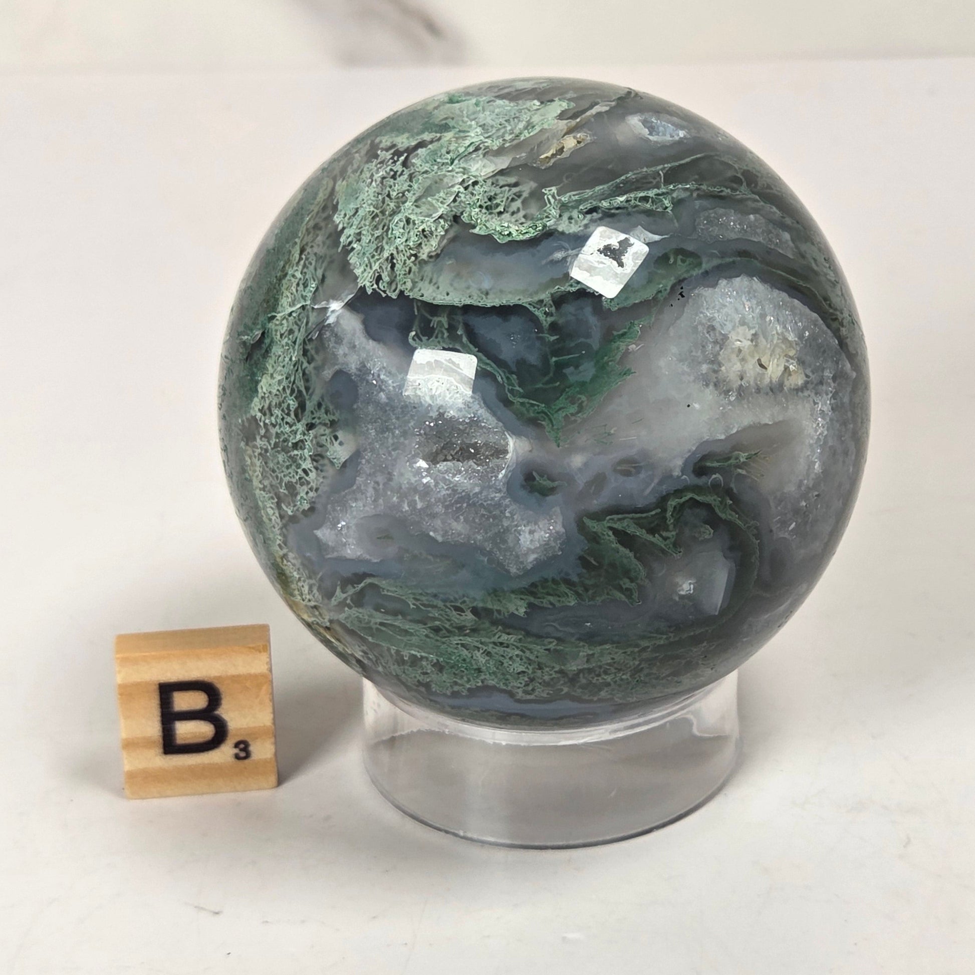 A polished geode-style decorative sphere with swirling green, gray, and white marbling, displayed on a clear stand next to a small wooden block letter tile on a light surface.