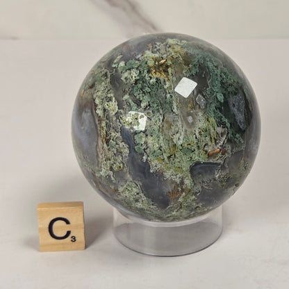 A polished natural stone sphere with mottled green, gray, and dark marbling, displayed on a clear stand next to a small wooden Scrabble tile showing the letter C.