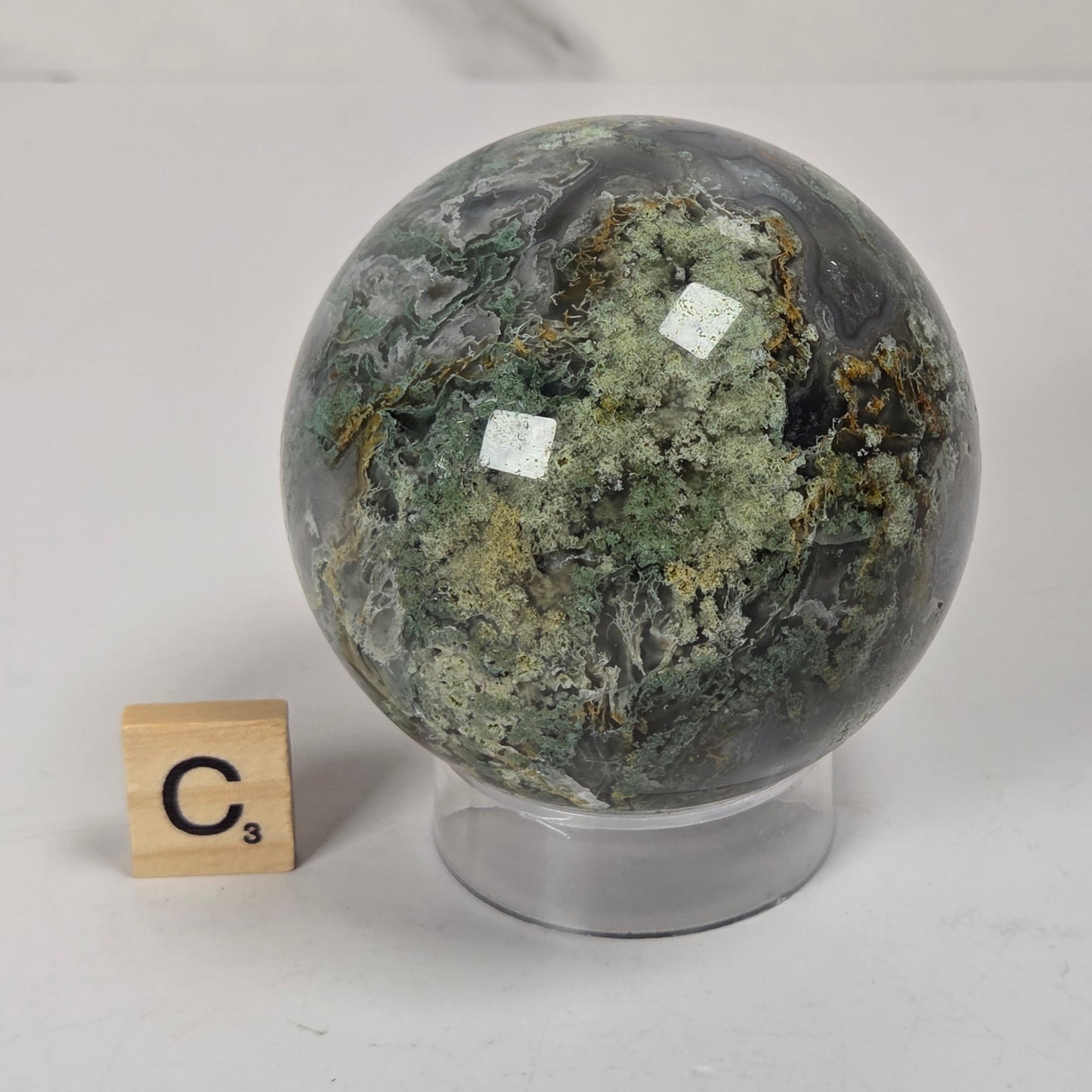 A polished green and gray banded stone sphere with natural mineral patterns, displayed on a clear acrylic stand next to a small wooden letter tile showing 'C3', set against a light gray marble-patterned background.