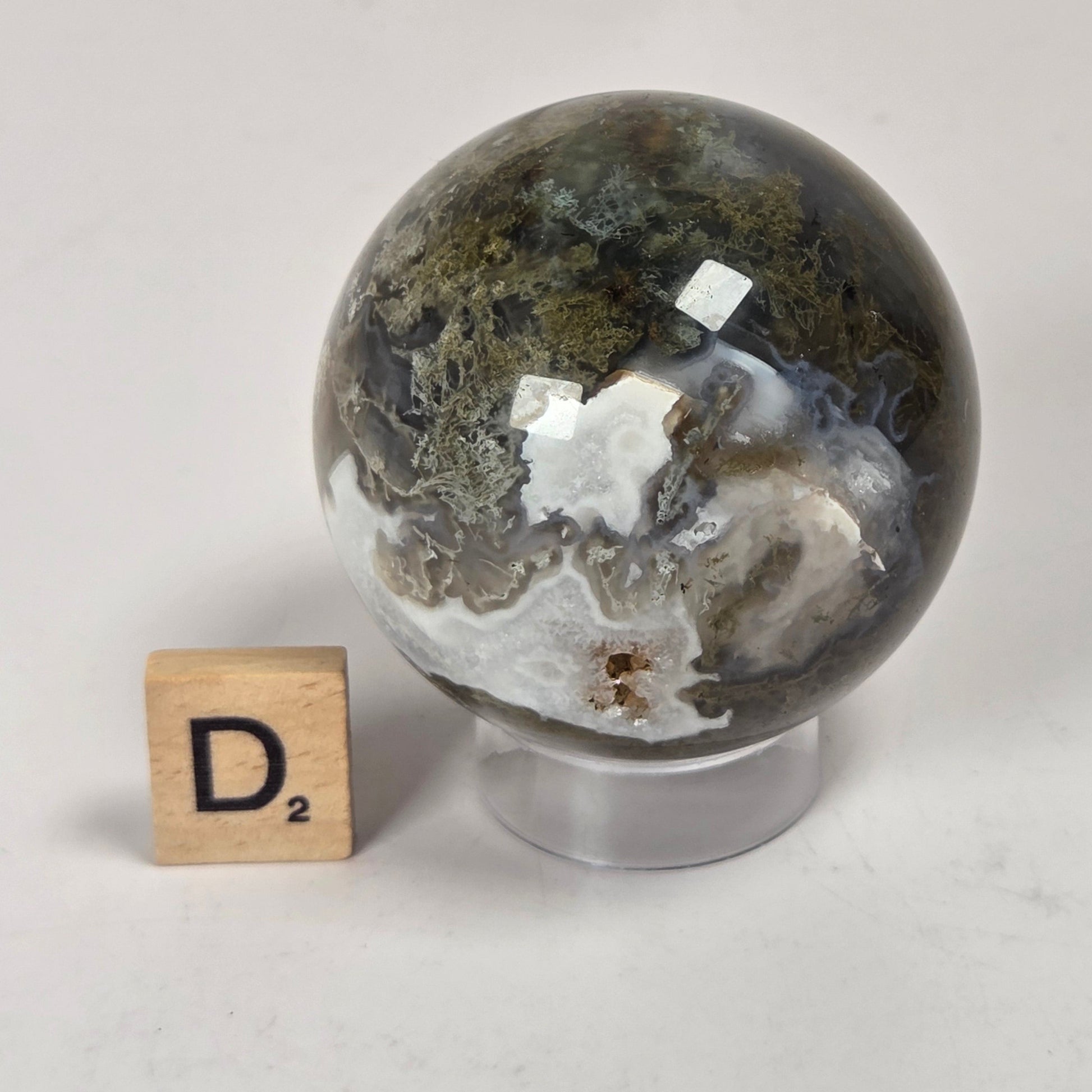 A polished round agate or marble-like crystal sphere on a clear acrylic stand, photographed on a light surface with a marble-pattern backdrop. A small wooden letter tile showing D² sits beside it for scale.