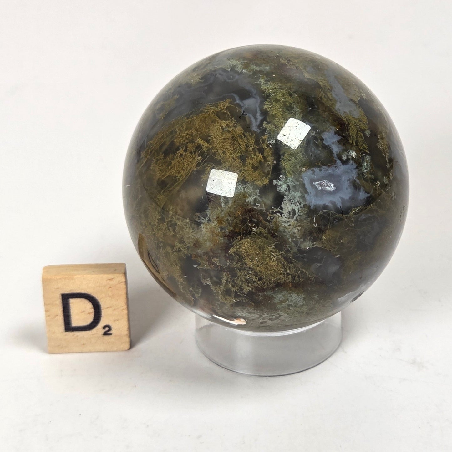 A polished dark green and black stone sphere with natural marbling sits on a clear acrylic stand, next to a wooden Scrabble tile showing the letter D2, on a white surface.
