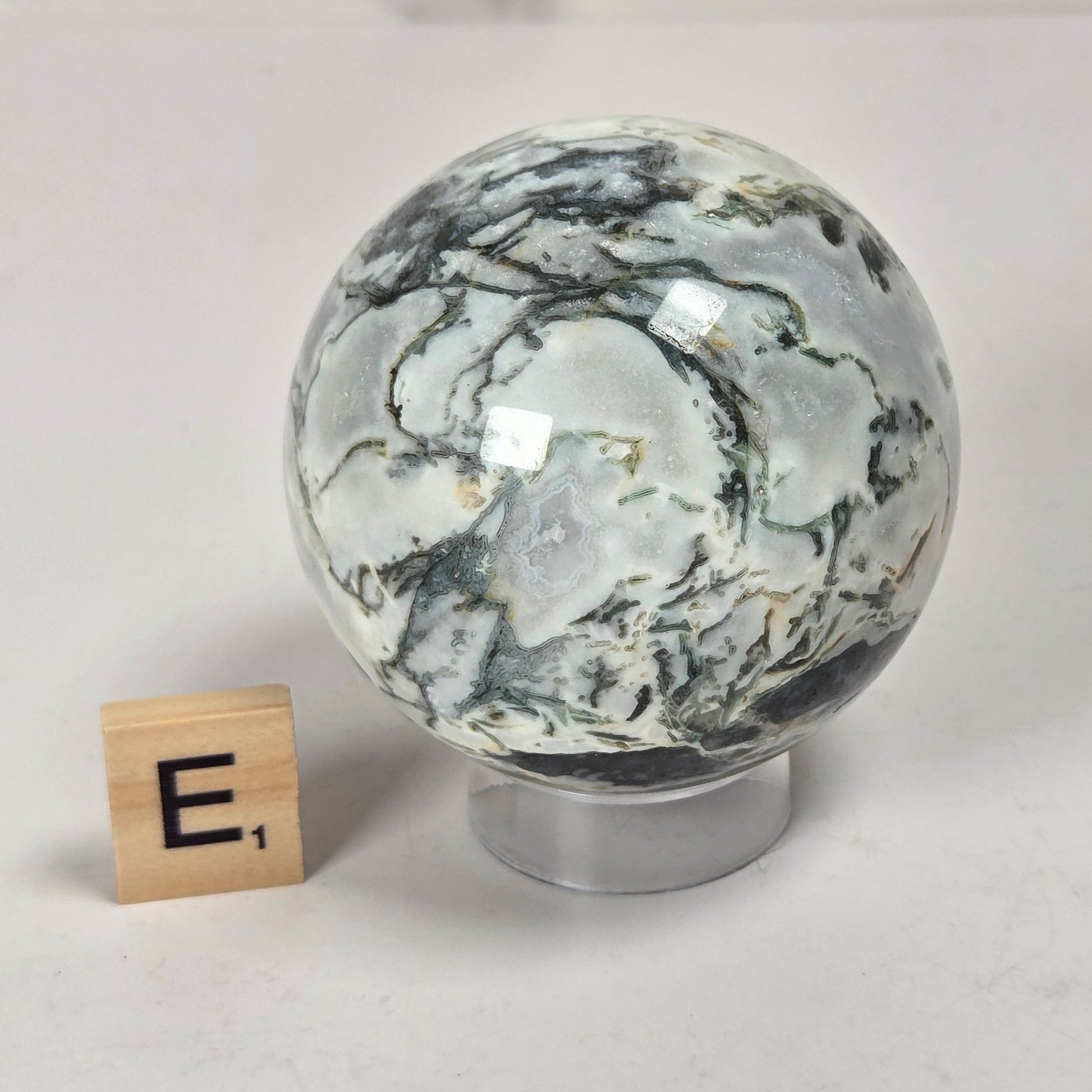 A polished marble-like decorative sphere with swirling white, gray, and dark charcoal veining, displayed on a small clear stand next to a wooden letter tile showing 'E1' on a light gray surface.