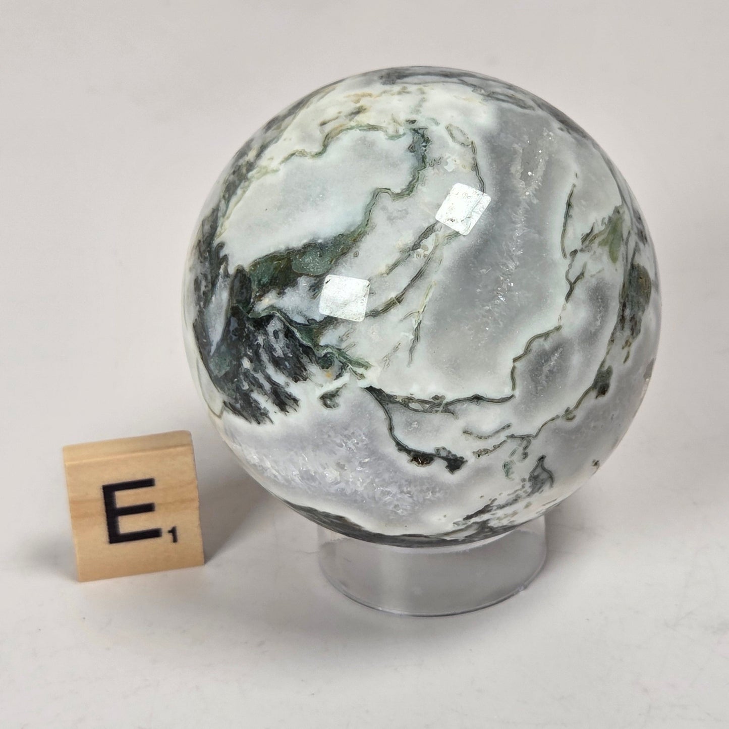 A polished marble-like decorative sphere with swirling white, gray, and dark green veining, displayed on a clear stand next to a small wooden block tile marked 'E1' on a light surface.