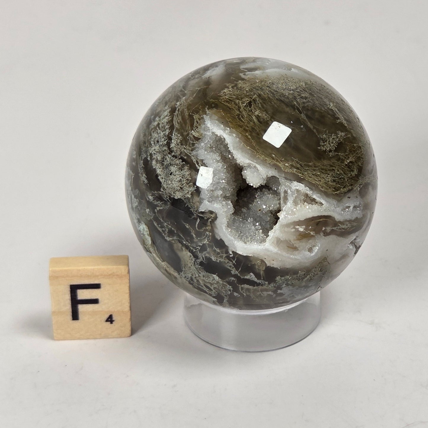 A polished natural stone sphere with swirling gray, olive-green, and white marbling, displayed on a clear acrylic stand next to a small wooden letter block with the letter F on a light gray surface.