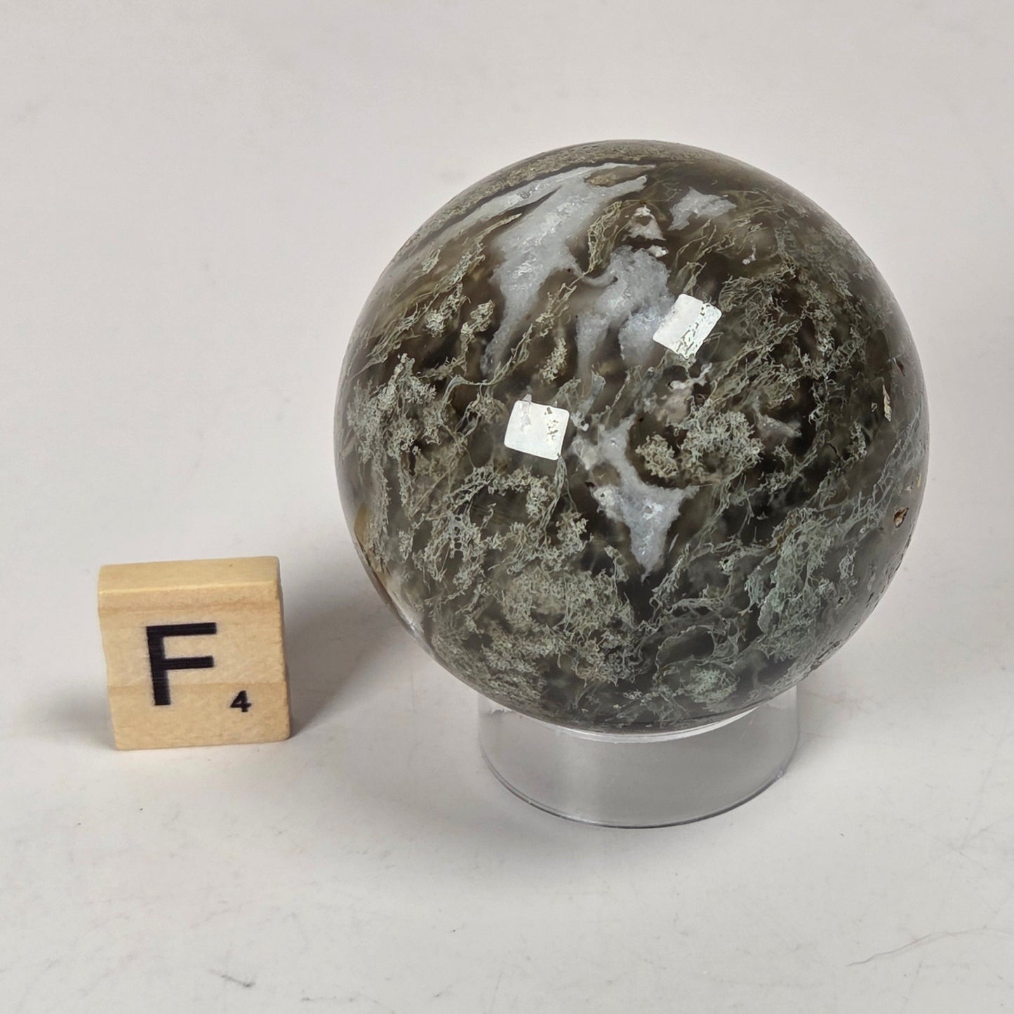 A polished natural stone sphere with dark gray, green, and white marbling, displayed on a clear acrylic stand next to a wooden Scrabble tile showing the letter F.
