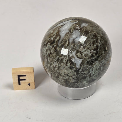 A polished natural stone sphere with dark gray, green, and white marbling, displayed on a clear acrylic stand next to a wooden Scrabble tile showing the letter F.