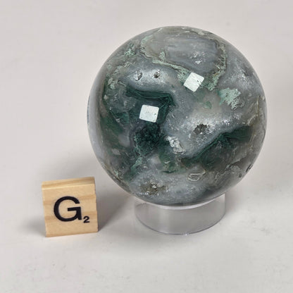 A polished round stone sphere with swirling dark green, gray, and white marbling, displayed on a clear stand next to a wooden Scrabble tile on a light gray surface.