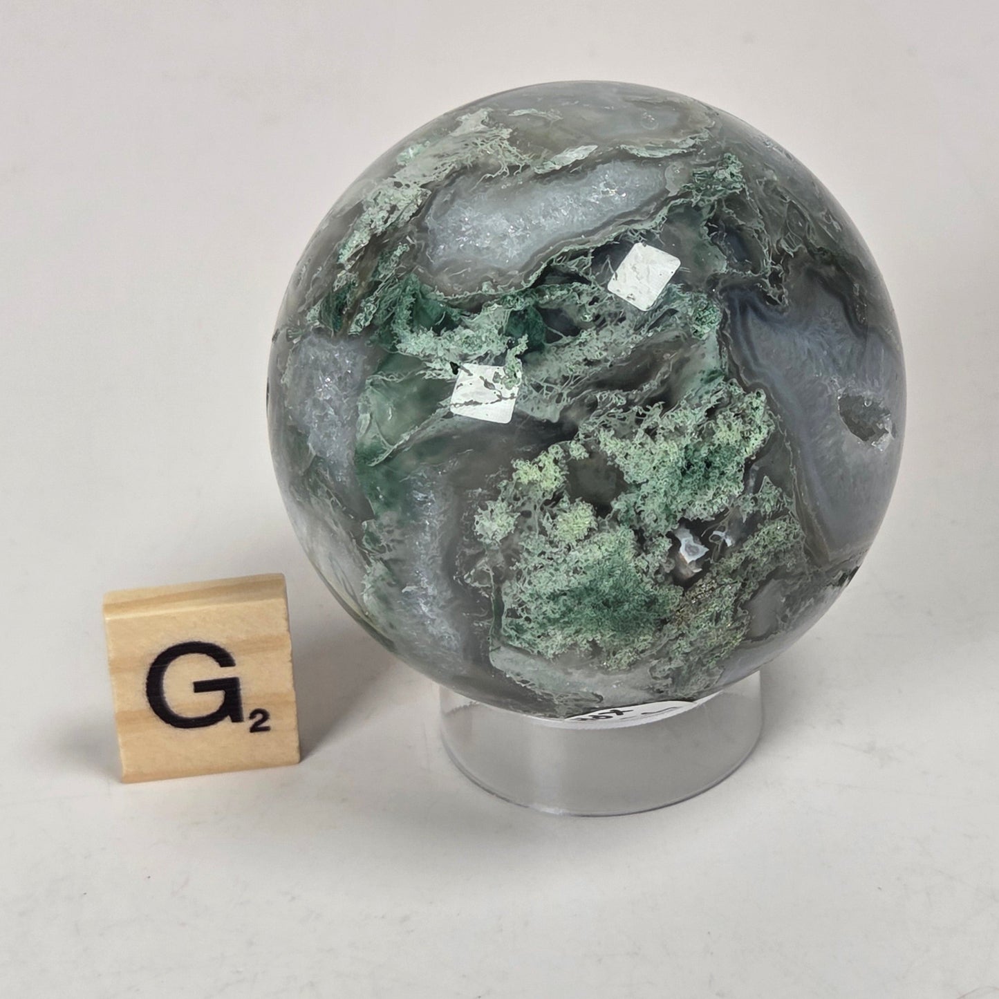 A polished round stone sphere with green and gray marbled patterns, displayed on a clear stand next to a small wooden letter tile spelling G2, on a light gray surface.